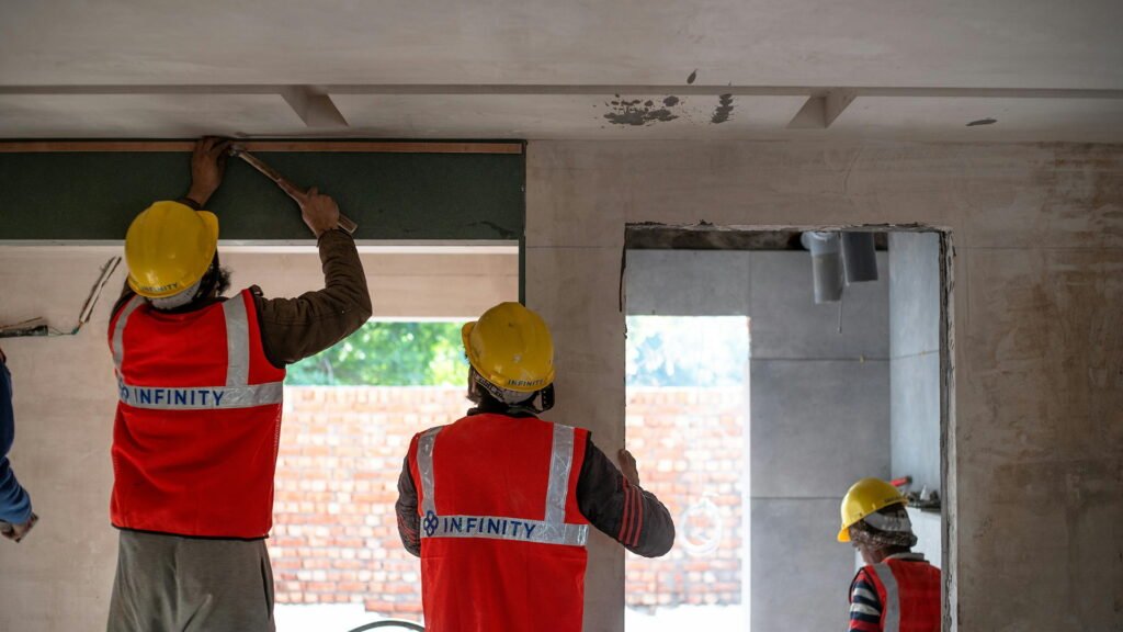A team of construction workers in helmets and reflective vests collaborate on installing panels within a partially built structure, framed by brick walls and greenery beyond.