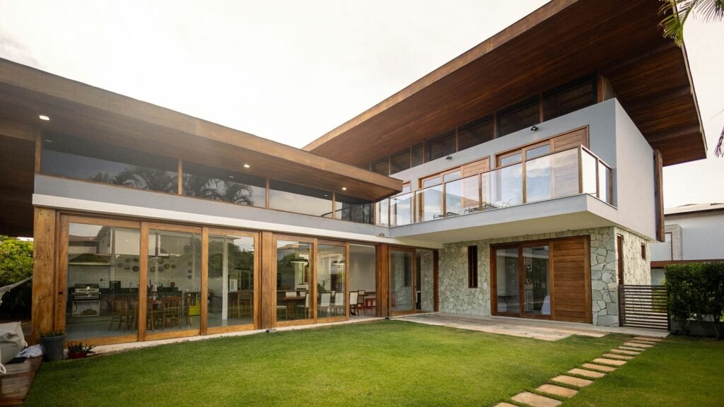 A sleek residence featuring expansive glass windows, warm wooden framing, and a stone façade, complemented by a manicured lawn and geometric overhang.