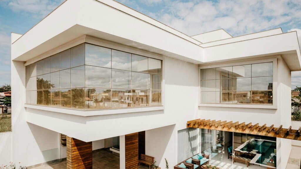 refined structure with clean lines, white walls, and wraparound glass, accented by a wooden pergola, outdoor seating, and a poolside view.