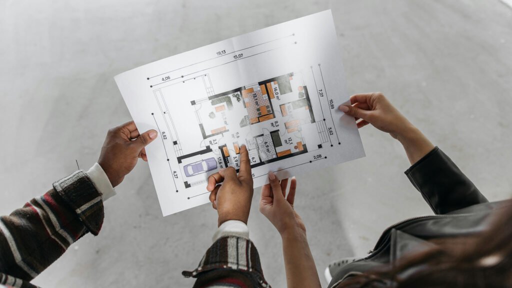 Close-up of hands holding and pointing at a printed architectural blueprint showing a modern home layout with labeled dimensions and furniture zones — symbolizing design collaboration and planning precision.