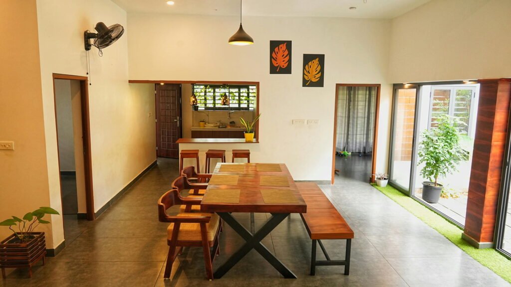 A bright, open-plan dining area with a solid wood table, woven chairs, and a bench, complemented by indoor plants and large glass doors — blending earthy tones with modern simplicity.